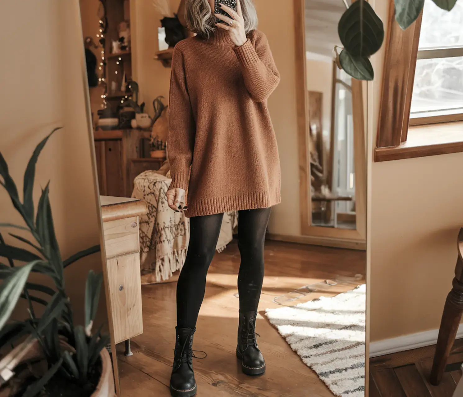 A woman takes a mirror selfie, wearing combat boots, a sweater dress, and tights; her face is not visible in the image
